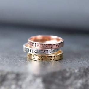 Pode incluir: Três anéis empilhados com coordenadas gravadas. O anel superior é de ouro rosa, o anel do meio é de prata e o anel inferior é de ouro. As coordenadas estão gravadas na parte exterior de cada anel.