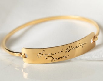 Puede incluir: Pulsera de brazalete de oro con una etiqueta rectangular curva grabada con el mensaje escrito a mano "Love & Blessings Mom".