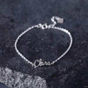 K&ouml;nnte beinhalten: Ein silbernes Kettenarmband mit dem Namen "Clara" in Kursivschrift auf einem silbernen Anh&auml;nger. Das Armband liegt auf einer dunkelgrauen, strukturierten Oberfl&auml;che.