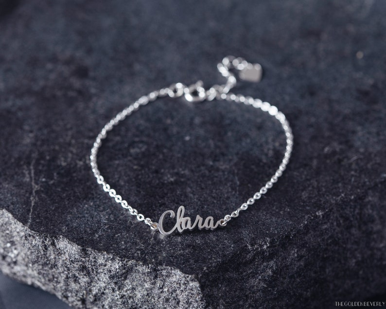 K&ouml;nnte beinhalten: Ein silbernes Kettenarmband mit dem Namen "Clara" in Kursivschrift auf einem silbernen Anh&auml;nger. Das Armband liegt auf einer dunkelgrauen, strukturierten Oberfl&auml;che.