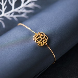 May include: A gold chain bracelet with a delicate monogram charm. The charm is made of intertwined letters, creating a stylish and personalized piece of jewelry.