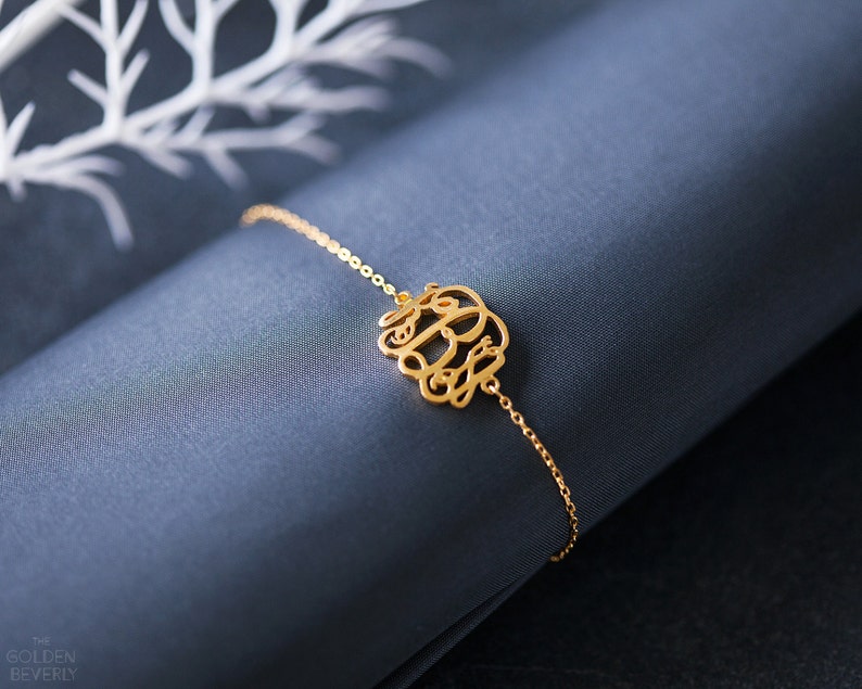May include: A gold chain bracelet with a delicate monogram charm. The charm is made of intertwined letters, creating a stylish and personalized piece of jewelry.