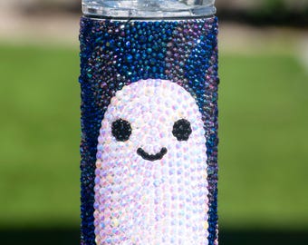 May include: A 15 oz rhinestone tumbler featuring a friendly ghost design. The tumbler is adorned with blue and purple rhinestones, and a white ghost with black eyes and a curved mouth. The text "FRIENDLY GHOSTS" and "15 OZ RHINESTONE TUMBLER" are at the top.