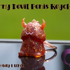 May include: A red and orange glitter resin keychain shaped like a devil with horns. The keychain is approximately 1 1/2 inches tall. The keychain is on a black surface with a pink and purple logo that says "kitten kreator".