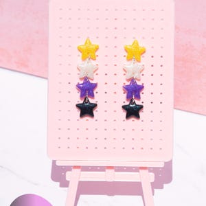 May include: Non Binary Pride Stud Earrings. Two dangling earrings featuring four stacked star-shaped charms in yellow, white, purple, and black glitter finishes. The earrings are displayed on a light pink pegboard stand with a kitten kreator logo.