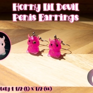 May include: A pair of pink, translucent devil-shaped earrings with black eyes and small horns, featuring metal hooks. The text reads: Horny Lil Devil Penis Earrings, and approximately 1 1/2 (t) x 1/2 (w). A circular inset shows the earrings glowing in the dark.