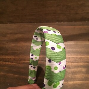 May include: A green and white headband with purple polka dots. The headband is made of woven ribbon.