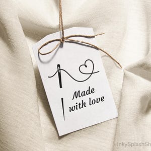 Puede incluir: Una etiqueta blanca con el texto "Made with love" y un dise&ntilde;o de aguja e hilo. La etiqueta est&aacute; atada con hilo y cuelga sobre un fondo de tela texturizada de color blanco roto.