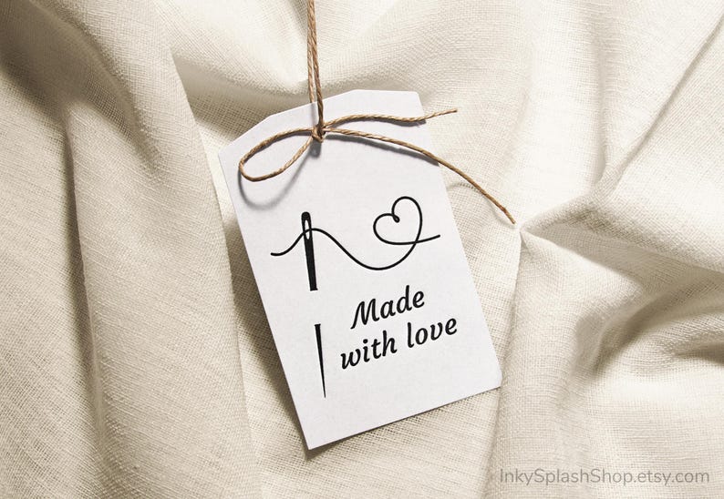 Puede incluir: Una etiqueta blanca con el texto "Made with love" y un dise&ntilde;o de aguja e hilo. La etiqueta est&aacute; atada con hilo y cuelga sobre un fondo de tela texturizada de color blanco roto.