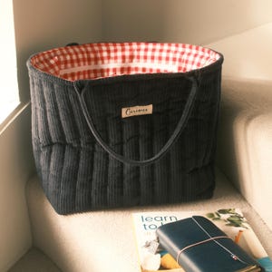 Può includere: Una borsa tote in velluto a coste grigio scuro con fodera a quadretti rossi e bianchi. La borsa ha una forma rettangolare con dettagli a coste verticali e due manici. Una piccola etichetta con la scritta "Curious" è cucita sul davanti.