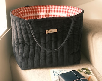 May include: A dark gray corduroy tote bag with a red and white gingham lining. The bag has a rectangular shape with vertical ribbed detailing and two handles. A small label that says "Curious" is sewn on the front.
