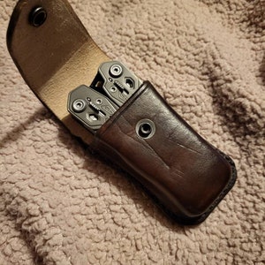 May include: A brown leather case with a snap closure, holding a multi-tool with a silver finish.