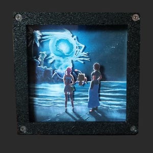 May include: A square shadow box art piece featuring a layered paper cutout scene of two figures and a small creature standing on a beach at night. The background displays a glowing blue moon over the ocean, framed by a textured black border.