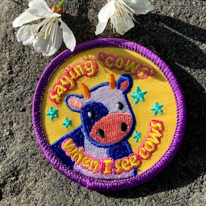 May include: Embroidered patch with a cartoon cow design. The patch features a yellow background with a purple border and the text "Saying Cows When I See Cows". The cow is blue and white with orange horns and pink nose. Green star accents.