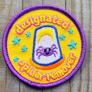 May include: A circular embroidered patch with a yellow background and a purple border. It features a white cup graphic with a purple spider inside, surrounded by light blue stars. The text reads designated spider remover in pink lettering.