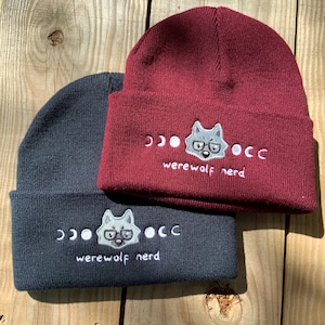 Op de afbeelding: Twee gebreide mutsen met een grijze wolf die een bril draagt en de tekst "werewolf nerd" geborduurd op de voorkant. Eén muts is bordeauxrood en de andere is donkergrijs.