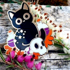 May include: A black cat enamel pin with gold edging, featuring a skull, potion bottles, and star details. The pin has orange, purple, and white colours. The pin is set against a backdrop of flowers and wood.