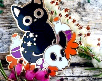 May include: A black cat enamel pin with gold accents, featuring a skull, potion bottles, and star details. The pin has orange, purple, and white colors. The pin is set against a backdrop of flowers and wood.