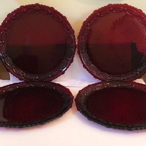 May include: Four round plates made of deep red pressed glass. Each plate features a central sunburst pattern radiating from the middle, surrounded by a wide, textured border with an embossed oval and bead design along the scalloped edge.