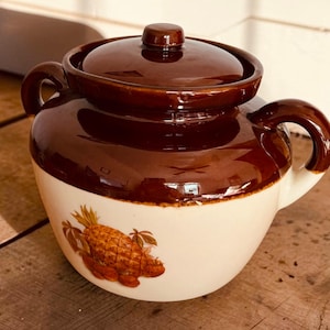 May include: A ceramic bean pot with a glossy dark brown lid and upper rim, featuring a cream-colored base. The pot has two side handles and a decorative pineapple illustration in shades of brown and orange on the front of the cream body.