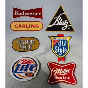 May include: A collection of vintage beer brand patches, including Budweiser, Blatz, Carling, Olympia Gold, Heileman's Old Style, Miller Lite, and Miller High Life. The patches feature various shapes and colors, with text in white and red.