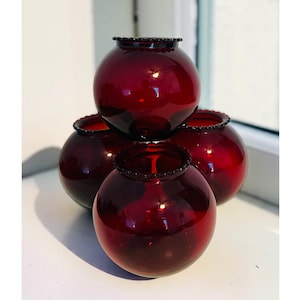 May include: A set of four spherical, deep red glass votive holders arranged in a pyramid formation. Each piece features a smooth, rounded body and a small, circular opening at the top with a delicate, beaded rim detail around the edge.