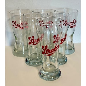 May include: Six clear glass beer glasses with a flared shape and a thick base. The glasses have the red text "Stroh's BEER" printed on them. The glasses are arranged in a semi-circle.