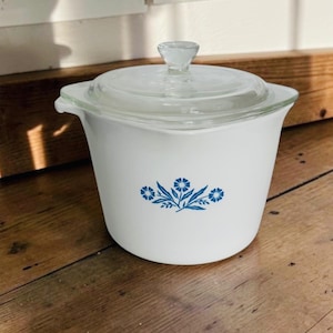 May include: A white glass casserole dish with a clear glass lid and a small knob handle. The exterior features a blue floral pattern consisting of three stylized flowers and leaves centered on the front. The vessel has a wide rim and a tapered base.