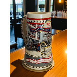 May include: A ceramic beer stein with a handle, featuring a detailed illustration of an American scene with a vintage car and the text "Great American Ale." The stein has a red, white, and blue color scheme, with a decorative rim and base.