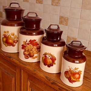 May include: Four ceramic kitchen canisters with lids. The canisters are cream-colored with brown lids and bases. Each canister features a different fruit design, including pears, grapes, and peaches.