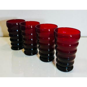 May include: Four ruby red drinking glasses with a ribbed design. The glasses have a dark base that fades into the vibrant red color at the top. The glasses are arranged in a row, showcasing their unique shape and color.