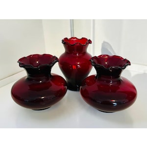 May include: Three deep red glass vases with ruffled, scalloped rims. Two vases feature a wide, rounded base and a short neck, while the central vase is taller with a more slender, bulbous body. All three pieces have a smooth, glossy finish.