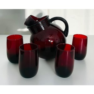 May include: A deep red glass pitcher with a curved handle and a matching set of four rounded, stemless drinking glasses. The glassware features a smooth, translucent finish with a consistent dark crimson hue throughout each individual piece.