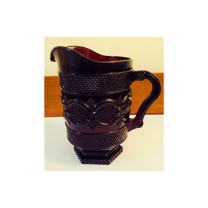 May include: A dark ruby-coloured glass jug with a textured surface and ornate detailing. The jug features a handle and a spout for pouring. The design includes diamond and floral patterns. The jug is likely vintage and suitable for serving beverages.