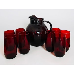 May include: A set of red glass drinkware, including a pitcher with a handle and spout, and several matching tumblers. The glassware is arranged on a white surface, creating a striking contrast.