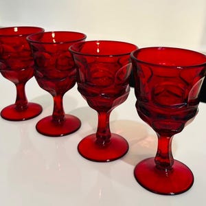 May include: A set of four matching ruby red glass goblets, each featuring a distinct circular embossed pattern on the bowl, a short stem, and a circular base. The glassware is translucent and uniform in color, showcasing a consistent design.