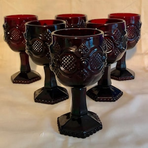 May include: Set of six ruby red goblets. Each goblet features an ornate floral and geometric design on the bowl and stem. The goblets have a hexagonal base and are made of glass. The goblets are arranged on a white surface.