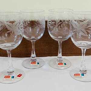 May include: Four clear wine glasses with etched floral designs. Each glass has a long stem and a round base. The etched design features leaves and other botanical elements. The glasses have a label that says "Fascination".