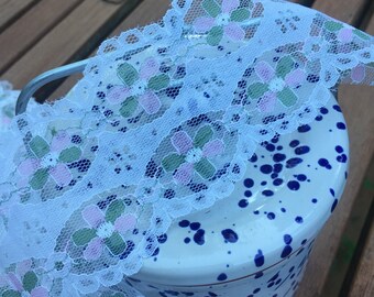 May include: White lace trim with pink and green floral embroidery. The lace is draped over a white ceramic jar with blue speckles.