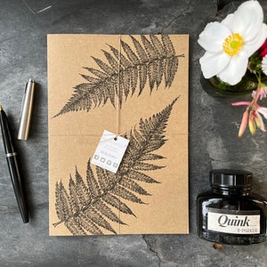 May include: A brown paper package featuring fern illustrations, tied with string. A black fountain pen and a silver pen cap are positioned to the left. A bottle of black ink and a small vase of flowers are on the right. The ink bottle has a white label that reads "Quink + PARKER".
