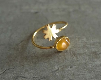 May include: A gold-toned open ring featuring a small, flat, six-petaled flower cutout on one end and a smooth, round, amber-colored cabochon stone set in a gold-toned bezel on the other end, connected by a thin, circular gold-toned metal band.
