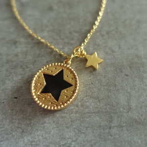 May include: Gold necklace with a black star charm and a smaller gold star charm. The necklace is on a gray background.