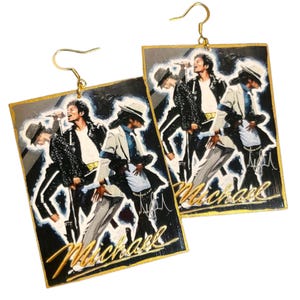 May include: A pair of rectangular drop earrings featuring a painted illustration of Michael Jackson in various performance poses. The design includes the name Michael in gold script at the bottom, set against a black background with gold edges.