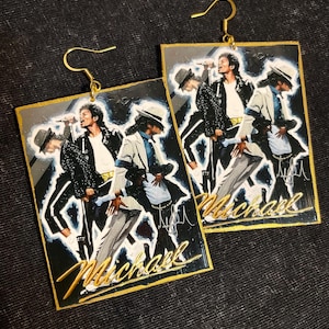 May include: A pair of rectangular earrings featuring a painted image of Michael Jackson in a performance pose. The earrings have a gold border and gold-colored fish hook ear wires. The word "Michael" is written in gold script across the bottom of each earring.