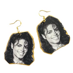 May include: A pair of dangle earrings featuring a black and white photographic portrait of Michael Jackson. The earrings have an irregular, rock-like shape with gold-colored edges and gold-toned metal hooks for attachment to the ear.