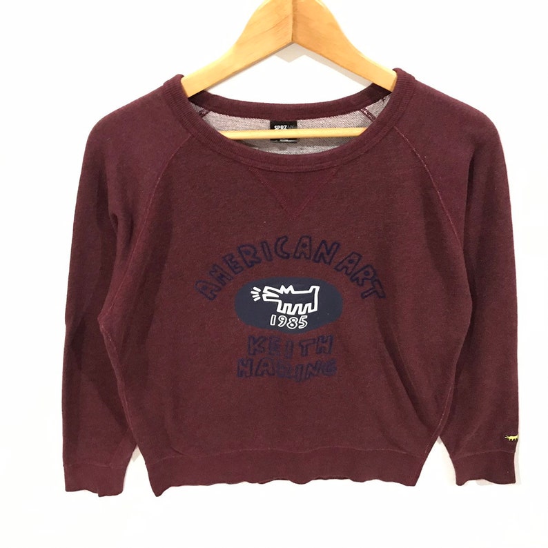 May include: Maroon sweatshirt featuring the text "AMERICAN ART" and "KEITH HARING 1985" in navy blue. The design includes a white dog-like figure inside a navy blue oval. The sweatshirt is displayed on a wooden hanger.