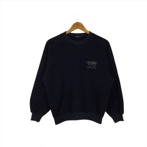 May include: A navy blue sweatshirt with a crew neck and long sleeves, hanging on a wooden hanger. The sweatshirt features a small, triangular logo with the text "ELLESSE" and "SINCE 1962" in a light color.