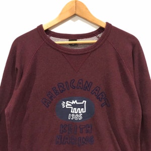 May include: A maroon sweatshirt featuring navy blue text that reads "AMERICAN ART". Below this is a navy blue oval with a white dog graphic and the number "1985". The name "KEITH HARING" is printed below the oval in navy blue.