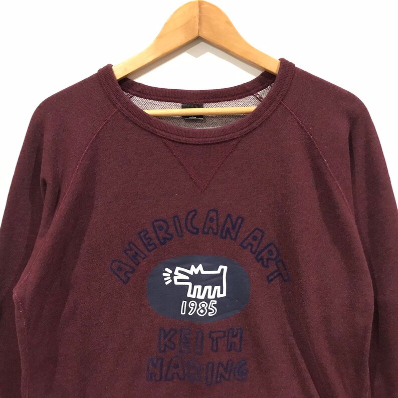 May include: A maroon sweatshirt featuring navy blue text that reads "AMERICAN ART". Below this is a navy blue oval with a white dog graphic and the number "1985". The name "KEITH HARING" is printed below the oval in navy blue.