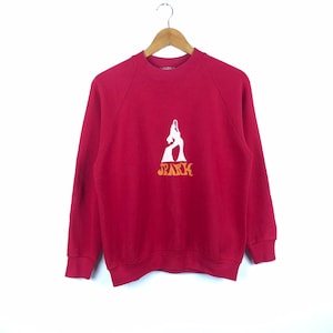 May include: A red sweatshirt with a white graphic of a figure and the word "SPARK" in orange. The sweatshirt has a crew neck and long sleeves. The garment is hanging on a wooden hanger.